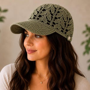May include: An olive green baseball cap crafted from crocheted yarn featuring an intricate openwork leaf pattern across the crown and a solid crocheted brim. A small, circular, light-colored button is visible on the side near the lower edge.