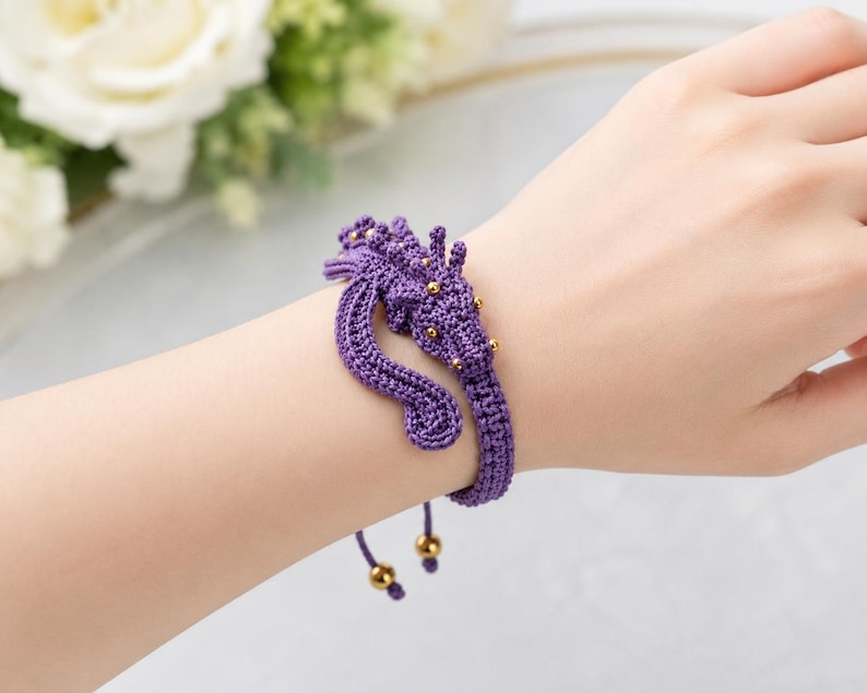 May include: A purple crocheted bracelet featuring a dragon-shaped centerpiece adorned with small gold-colored beads. The bracelet has a woven band and two dangling cords, each finished with a single gold-colored bead at the end for adjustment.
