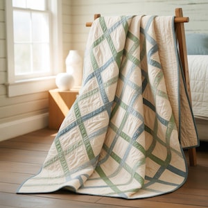 May include: A cream-coloured quilt with a grid pattern in shades of blue and green is draped over a wooden rack. The quilt has a blue border and is displayed in a well-lit room with a window and wooden floor.