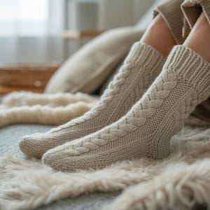 May include: A pair of beige cable-knit socks. The socks are knee-high and feature a braided design along the front. They are resting on a soft, cream-colored blanket. The socks appear warm and cozy.