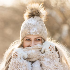 May include: A woman bundled in winter wear, including a beige knit hat with a fluffy pom-pom, a matching scarf, and gloves. The gloves have a snowflake design. Snowflakes are visible on the hat and coat.