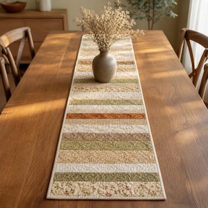 May include: A quilted table runner with a patchwork design in shades of beige, green, and brown. The runner features floral and paisley patterns, and is placed on a wooden table. A vase with dried flowers sits on top of the runner.