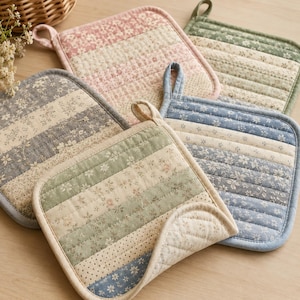 May include: A collection of five square quilted pot holders featuring horizontal stripes with various floral and dotted patterns in muted shades of pink, sage green, blue, and cream, each equipped with a fabric hanging loop at the corner.