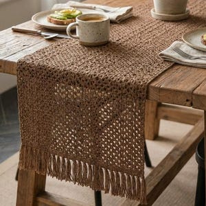 Może przedstawiać: Rustykalny drewniany stół jest nakryty brązowym, tkanym bieżnikiem z frędzlami. Na stole ustawiono dwa talerze z tostami z awokado, nakrapiany kubek i roślinę doniczkową. Scena skąpana jest w naturalnym świetle.