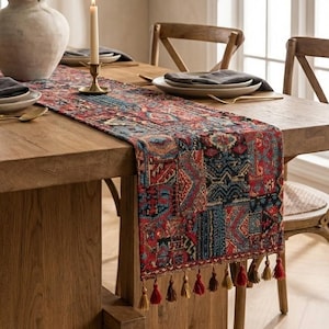 Op de afbeelding: Een houten eettafel met een patroon loper, borden en bestek. Een vaas met groen en brandende kaarsen dragen bij aan de inrichting. De loper heeft een rood, blauw en beige ontwerp met kwastjes.