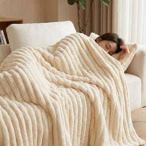 Puede incluir: Una persona está durmiendo en un sofá blanco, cubierta por una manta de felpa acanalada de color crema. La manta tiene un aspecto suave y texturizado. La escena está iluminada por la luz natural de una ventana cercana, creando un ambiente acogedor.