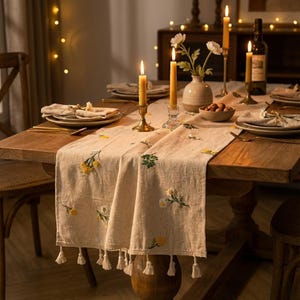 Op de afbeelding: Een houten eettafel gedekt voor een maaltijd, met een beige geborduurde tafelloper met gele en groene bloemmotieven. Aangestoken kaarsen, borden en een vaas met bloemen completeren de setting. De loper heeft witte kwastjes.