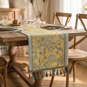 Puede incluir: Camino de mesa amarillo con estampado de cachemira, borde azul claro y borlas decorativas. El camino de mesa está colocado sobre una mesa de madera preparada para una comida, con platos, vasos y pan. La mesa está rodeada de sillas de madera.