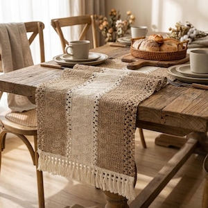 Może przedstawiać: Rustykalny drewniany stół jest nakryty tkanym bieżnikiem. Bieżnik ma kremowy środkowy panel otoczony brązowymi sekcjami z frędzlami. Na stole znajdują się talerze, kubki i kosz z chlebem.