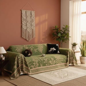 Puede incluir: Un sofá verde con una manta estampada con motivos botánicos y cojines decorativos. Un tapiz de macramé y plantas en macetas complementan la decoración de la habitación. Una pequeña alfombra y una mesa auxiliar.