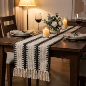 Może przedstawiać: Tkany bieżnik na stół z kremowym tłem i czarnymi geometrycznymi wzorami. Bieżnik ma frędzle na obu końcach. Stół jest nakryty talerzami, serwetkami, świecami, kwiatami i kieliszkami do wina.