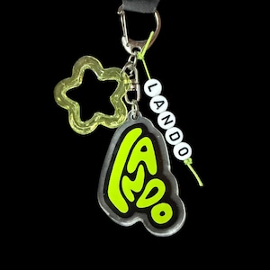 May include: A keychain with a black strap and silver clasp. The keychain features a clear, neon green star-shaped charm, a clear charm with the word "Lando" in neon green, and a beaded word "Lando". The background is black.