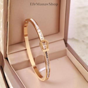 Könnte beinhalten: Ein goldfarbenes Armreif-Armband mit Schnallendesign, besetzt mit klaren Edelsteinen. Das Armband wird in einer Schmuckschatulle mit neutralem Innenraum präsentiert. Der Text "EfeManavShop" ist oben sichtbar.