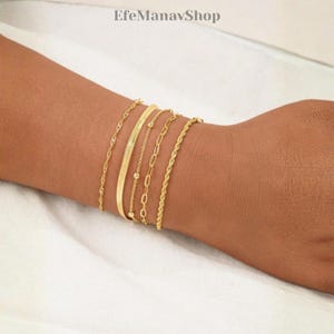 Puede incluir: Una colección de pulseras doradas en varios estilos, incluyendo un diseño de espiga, eslabones de cadena y cuerda. Las pulseras se muestran en el brazo de una persona, mostrando sus diferentes texturas y brillo. Las pulseras son de color oro cálido.