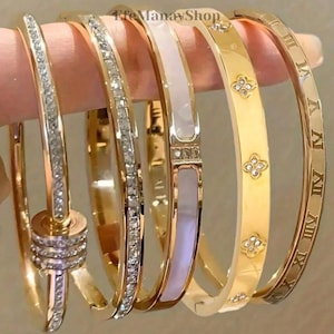 Può includere: Una collezione di braccialetti dorati, ognuno con design unici. Alcuni sono adornati con cristalli trasparenti, altri con motivi floreali, e uno presenta numeri romani. I braccialetti sono tenuti insieme, mostrando la loro varietà.