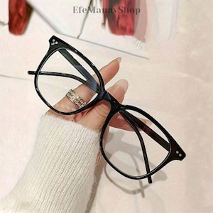 Op de afbeelding: Zwarte bril met een vierkante vorm. De bril heeft heldere glazen en wordt vastgehouden door een hand met een ring. De afbeelding heeft de tekst "EfeManavShop" linksboven.