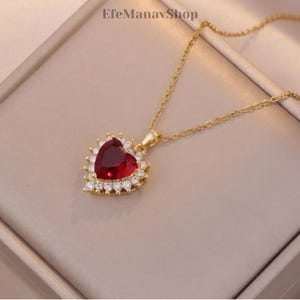 Puede incluir: Collar dorado con un colgante en forma de corazón con una gran gema roja oscura en el centro. El corazón está rodeado por un halo de gemas más pequeñas y claras. La cadena del collar es delicada y el colgante cuelga de una pequeña argolla.