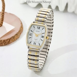 May include: A rectangular wristwatch with a white face, gold hands, and Roman numeral hour markers. The watch has a silver and gold-toned stretch band. The text "XAPWV" is printed on the watch face. The brand name "EfeManavShop" is visible in the top left corner.