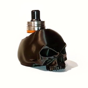 Può includere: Un accessorio per vaporizzatore a forma di teschio nero con un atomizzatore argentato e nero. Il teschio presenta dettagli ed è posto su uno sfondo bianco. L'atomizzatore ha un bocchino nero.