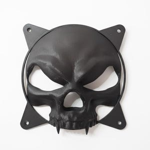 May include: Black skull-shaped decorative piece with a menacing expression. The skull has sharp teeth and is surrounded by a circular frame with four triangular mounting points. The piece is made of a matte black material.