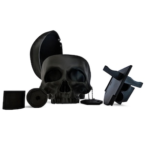 May include: A black skull-shaped container with a hinged lid, various accessories, and a small chain. The skull has detailed facial features and is surrounded by other black objects, including a stand and a cylindrical piece.