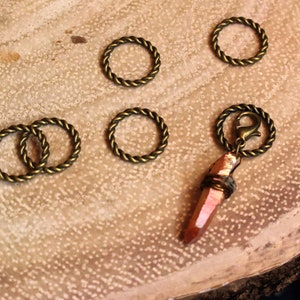 May include: A collection of gold-toned metal rings with a twisted rope design, arranged on a wooden surface. A single ring is attached to a long, pointed, orange crystal pendant.