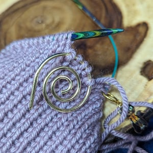 May include: A close-up of a purple knitted fabric with a gold spiral stitch marker. The fabric is being worked on with a pair of knitting needles.