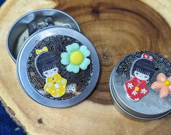 May include: Two round metal tins with lids. Each tin features a colorful illustration of a girl in a traditional Japanese kimono with a flower. One girl wears a yellow kimono with a green flower, and the other wears a red kimono with a pink flower.