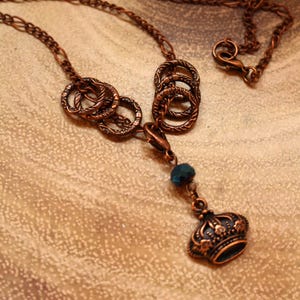 May include: A copper necklace with a chain and a pendant featuring a crown and a dark blue bead. The pendant is made of multiple interconnected rings.