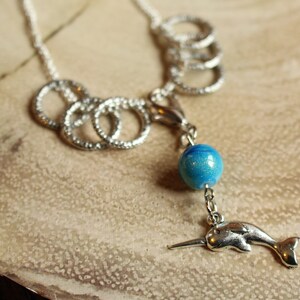 May include: A silver necklace with a blue and silver charm featuring a narwhal. The necklace has multiple silver rings attached to it.