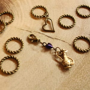 May include: A set of gold-toned metal ring stitch markers with a heart charm and a teapot charm. The charms are attached to a chain with a purple bead.