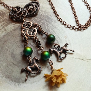 May include: A copper necklace with a detailed pendant featuring a floral design and three horse charms. The pendant is accented with iridescent green beads and a small yellow flower charm.