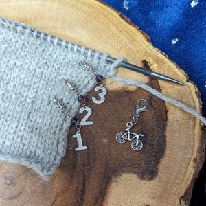 May include: Gray knitted fabric with three stitch markers with numbers 1, 2, and 3. A silver bike charm is attached to a stitch marker.
