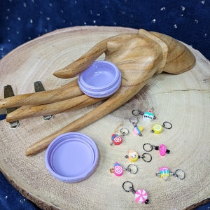 May include: A set of 10 colorful stitch markers shaped like different desserts, including a cupcake, ice cream cone, cake slice, and lollipop. The markers are stored in a small purple container with a lid.