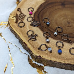 May include: A collection of 11 copper stitch markers with various charms, including a crown, a horse, a pine cone, and a variety of colored beads. The markers are displayed on a wooden surface.