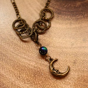 May include: A bronze necklace with a crescent moon charm and a dark iridescent bead. The necklace features a chain with multiple linked circles.