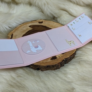 May include: A pink and white notepad with a llama design. The notepad has a flap that folds over to reveal a smaller notepad with a giraffe design. The notepad is on a wooden surface.