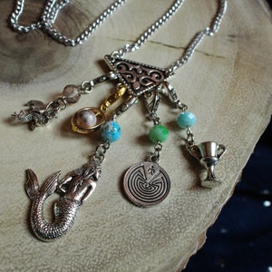 May include: A silver necklace with a triangular pendant and multiple charms, including a mermaid, a maze, a goblet, and a seahorse. The charms are attached to the pendant with silver chains and beads.