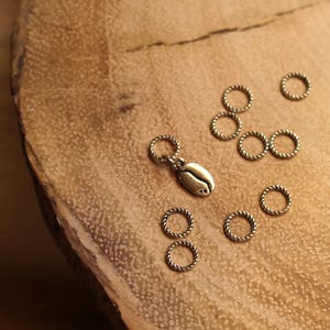 May include: A set of silver-toned metal jump rings with a coffee bean charm. The jump rings are arranged on a wooden surface.