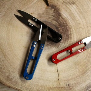 May include: Three pairs of small thread snips with different colored handles. The snips are on a wooden surface. The snips are black, blue, and red.