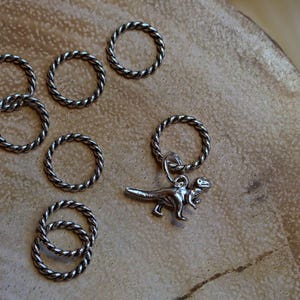 Może przedstawiać: Zestaw srebrnych, skręconych metalowych pierścieni, z jednym pierścieniem trzymającym srebrny wisiork w kształcie dinozaura. Pierścienie są ułożone na jasnobrązowej drewnianej powierzchni.