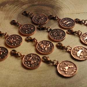 May include: Twelve copper-colored charms with zodiac symbols. Each charm has a loop for attaching to a chain or bracelet. The charms are arranged on a wooden surface.