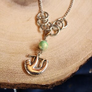 May include: A silver necklace with a silver sloth charm and a green gemstone bead. The necklace has a chain link design with multiple silver rings.