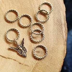 May include: A set of silver-toned metal stitch markers with a twisted rope design. One marker features a small, silver-toned fairy charm.