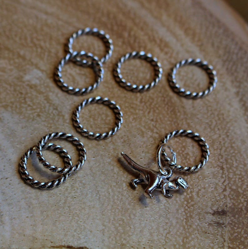 Może przedstawiać: Zestaw srebrnych, skręconych metalowych pierścieni, niekt&oacute;re ułożone jeden na drugim, oraz srebrny zawieszka w kształcie dinozaura. Pierścienie są używane do robienia na szydełku.