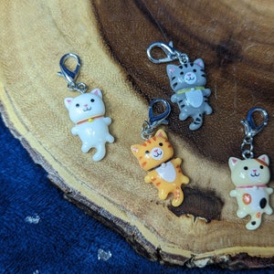 May include: Four colorful cat charm zipper pulls. The charms are made of plastic and feature a white cat, a gray cat, an orange cat, and a white cat with orange and black spots. Each charm has a silver metal zipper pull.
