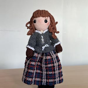 May include: A handmade crochet doll featuring brown yarn hair, a gray jacket with white lace-up detailing and white trim, and a long skirt with a blue, red, and beige plaid pattern. The doll has black bead eyes and stands on a small circular base.