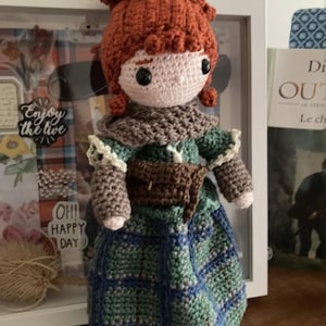 Puede incluir: Muñeca de ganchillo hecha a mano con cabello rojo y un vestido a cuadros verde y azul. La muñeca tiene un bolso marrón y una bufanda. El cabello de la muñeca está peinado de una manera única y texturizada. La muñeca es un personaje de una historia.