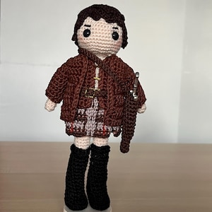 Könnte beinhalten: Eine handgefertigte Häkelpuppe mit dunkelbraunem Haar, schwarzen Perlenaugen und heller Haut. Die Puppe trägt eine braune Jacke über einem karierten Rock, einen braunen Gürtel und hohe schwarze Stiefel. Ein kleines silberfarbenes Schwert hängt am Riemen.