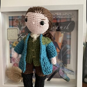 May include: A handmade crochet doll with brown hair and black eyes, wearing a green vest, blue jacket, brown shorts, and black boots. The doll is displayed in front of a framed background with decorative elements.